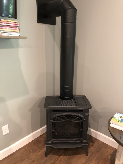 A black wood stove with a vertical chimney pipe, positioned against a light gray wall.