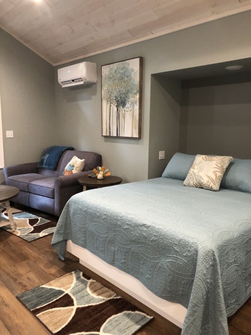 Cozy bedroom with a blue bedspread, a gray sofa, and a wall art of trees, featuring wooden flooring and decorative rugs.