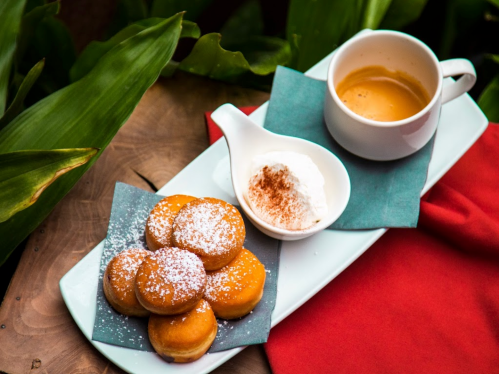 A plate with six powdered sugar-dusted pastries, a small bowl of cream, and a cup of espresso on a wooden surface.