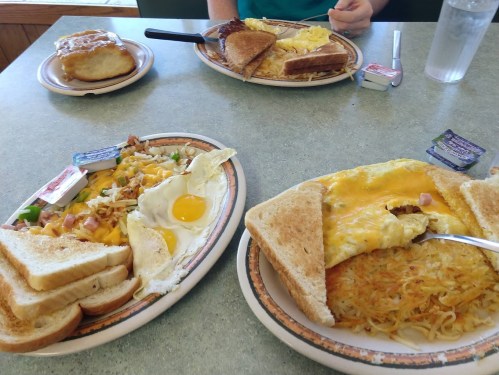 A table with three breakfast plates: eggs, toast, hash browns, and a biscuit with gravy in the background.