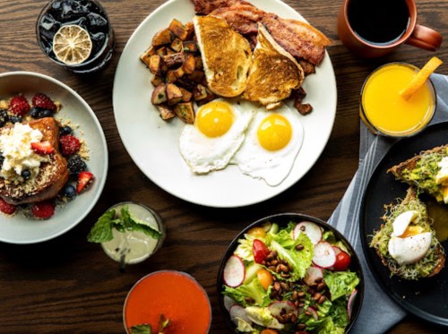 A spread of breakfast dishes including eggs, bacon, toast, fruit, salad, and various drinks on a wooden table.