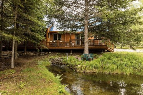 A cozy wooden cabin surrounded by trees, with a small stream and green foliage in the foreground.