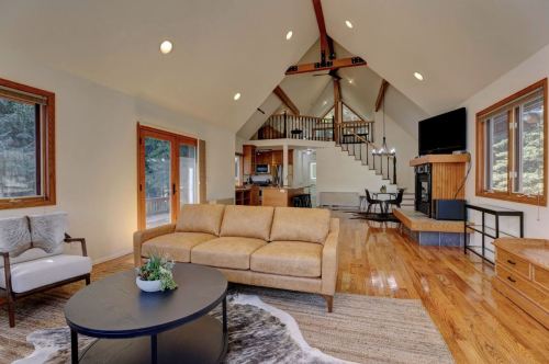 Cozy living room with a high ceiling, wooden floors, a sofa, and large windows overlooking a forested area.