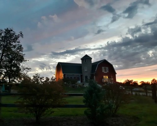 A red barn with a tower sits against a colorful sunset, surrounded by trees and a grassy field.