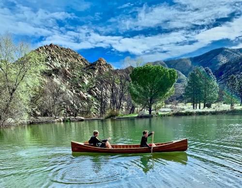 Two people paddle a wooden canoe on a calm lake surrounded by green trees and rocky hills under a blue sky.