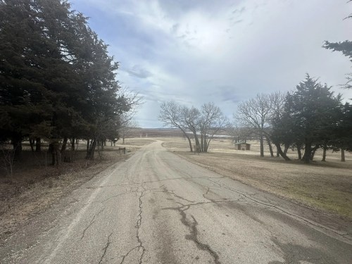 A cracked road winds through a rural landscape with sparse trees and a cloudy sky in the background.