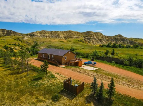 Aerial view of a brown house on a dirt road, surrounded by green hills and trees under a partly cloudy sky.