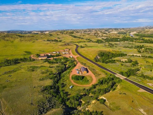 Aerial view of a rural landscape with rolling hills, a winding road, and scattered buildings amidst green fields.