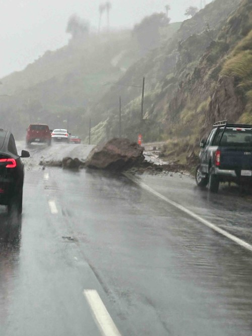 A large rockslide blocks a wet road during heavy rain, with cars navigating around the debris.