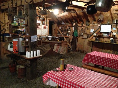 A rustic barn interior with wooden beams, checkered tablecloths, and various kitchen utensils hanging on the walls.