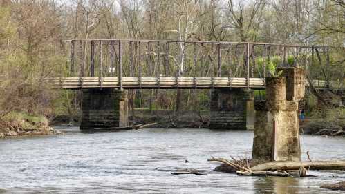 A weathered metal bridge spans a river, surrounded by trees and greenery, with a stone pillar visible in the water.