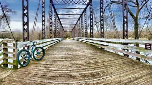 A bicycle rests on a wooden bridge with metal supports, surrounded by trees and a calm river.