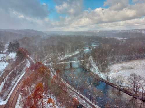 A snowy landscape with a river, trees, and a bridge, under a cloudy sky. Snowflakes are gently falling.