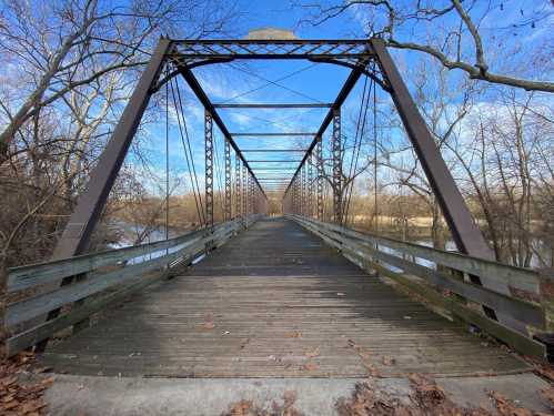 A wooden bridge with metal supports stretches over a river, surrounded by bare trees and a clear blue sky.