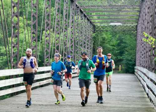 A group of six runners crosses a wooden bridge surrounded by greenery, enjoying a sunny day outdoors.