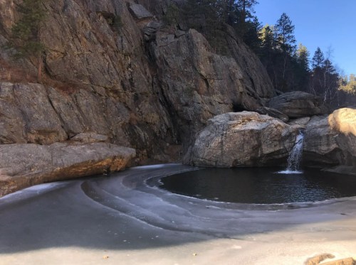 A serene landscape featuring a frozen pond surrounded by rocky cliffs and a small waterfall. Pine trees in the background.