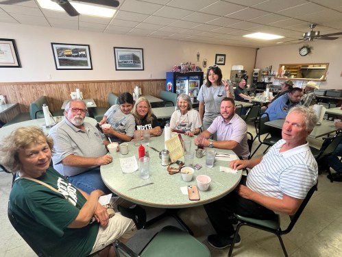 A group of eight people sitting around a table in a diner, smiling and enjoying their time together.