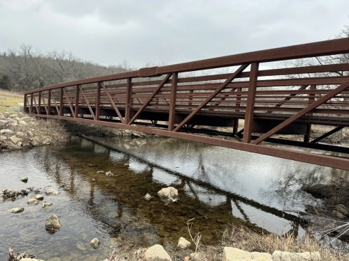 A metal bridge spans a shallow stream, surrounded by rocky banks and bare trees under a cloudy sky.