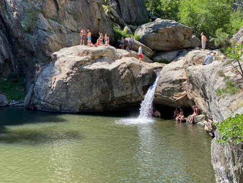 A scenic rocky area with a small waterfall flowing into a pool, where people are swimming and relaxing on the rocks.