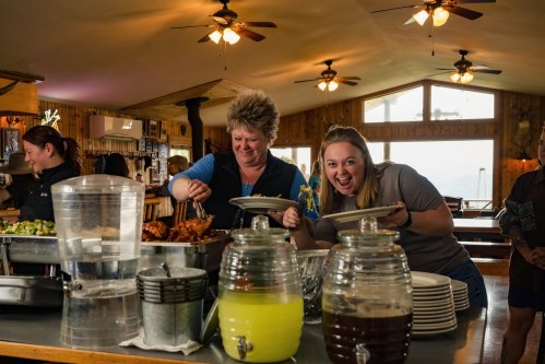 Two women smile while serving food at a buffet in a cozy, rustic dining area with wooden decor.