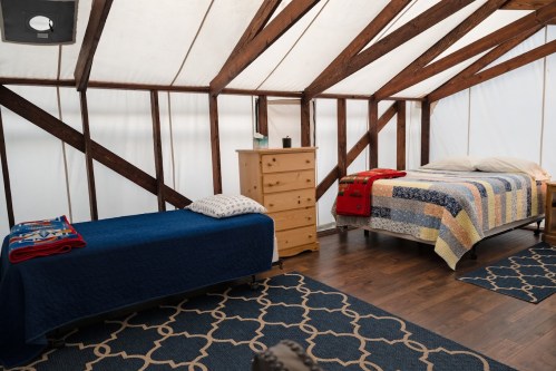 A cozy bedroom with two beds, a wooden dresser, and patterned rugs, set in a bright, rustic space with wooden beams.