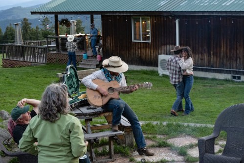 A cowboy plays guitar while couples dance outdoors near a rustic cabin in a scenic setting.