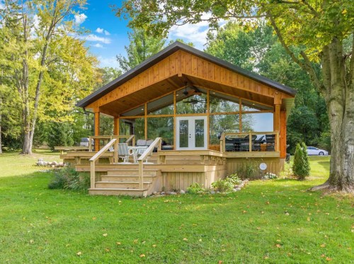 A modern wooden cabin with a large deck, surrounded by greenery and trees, under a clear blue sky.