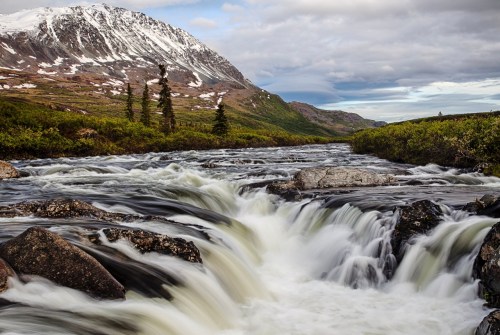 A rushing river flows over rocks, surrounded by lush greenery and mountains under a cloudy sky. Snow caps the peaks.
