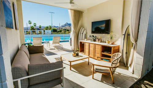 A stylish outdoor lounge area with a sofa, coffee table, and surfboards, overlooking a pool and mountains.