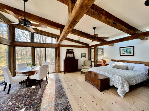Cozy bedroom with wooden beams, a large bed, dining area, and windows overlooking a forested view.