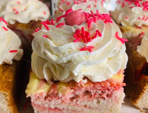 Close-up of a slice of cake topped with whipped cream and pink sprinkles, showcasing layers of pink and yellow cake.