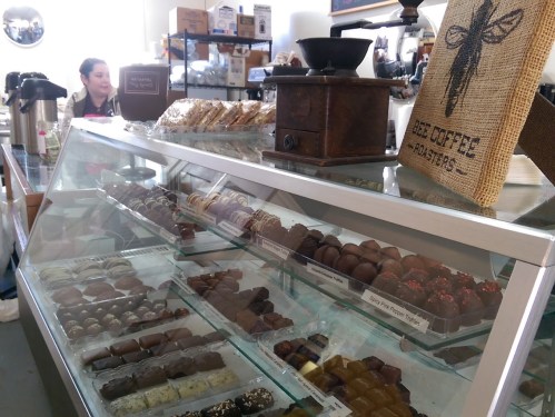 A display case filled with assorted chocolates and treats, with a barista in the background at a cozy café.
