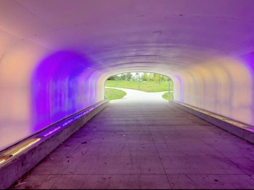 A brightly lit tunnel with purple hues, leading to a grassy area and pathway outside.