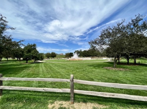 A grassy park with a wooden fence, trees, and a fountain under a blue sky with scattered clouds.