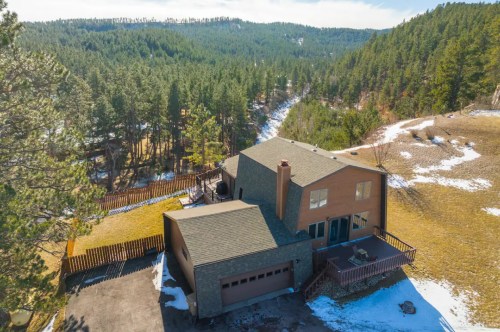 Aerial view of a spacious house surrounded by trees and hills, with a driveway and snow patches in the landscape.