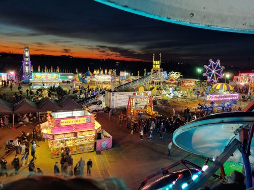 A vibrant carnival scene at dusk, featuring rides, food stalls, and colorful lights against a sunset sky.