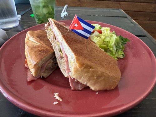 A Cuban sandwich with ham and cheese, garnished with a small Cuban flag, served with a side of lettuce on a red plate.