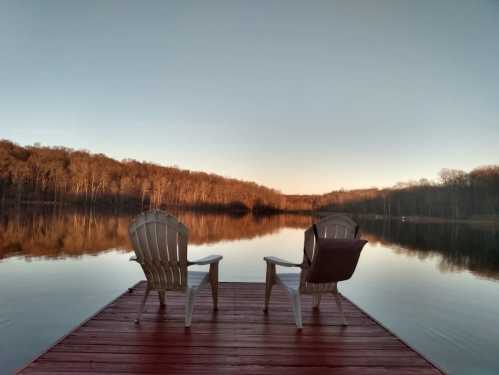 Two white chairs on a wooden dock overlooking a calm lake surrounded by trees at sunset.