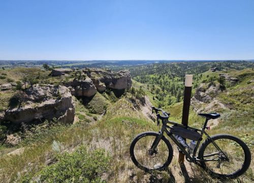 A bicycle rests near a trail marker, overlooking a scenic valley with green hills and clear blue skies.