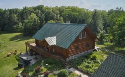 Aerial view of a rustic log cabin with a green metal roof, surrounded by trees and a landscaped yard.