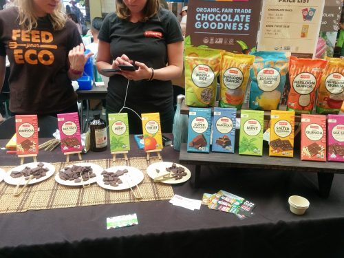 A colorful display of chocolate bars and snacks at a market booth, with people sampling and checking their phones.