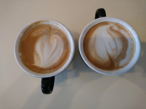 Two cups of coffee with latte art, featuring swirls and heart shapes, placed on a light-colored surface.