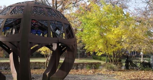 A large, dark metal globe sculpture surrounded by autumn trees with yellow leaves in a park setting.