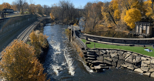 A river flows past a park with autumn trees, rocky banks, and a nearby railway track under a clear blue sky.