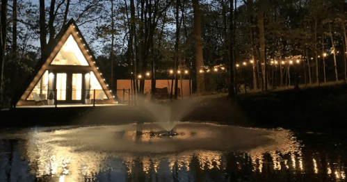 A triangular cabin by a pond, illuminated by string lights, with a fountain creating ripples in the water at dusk.