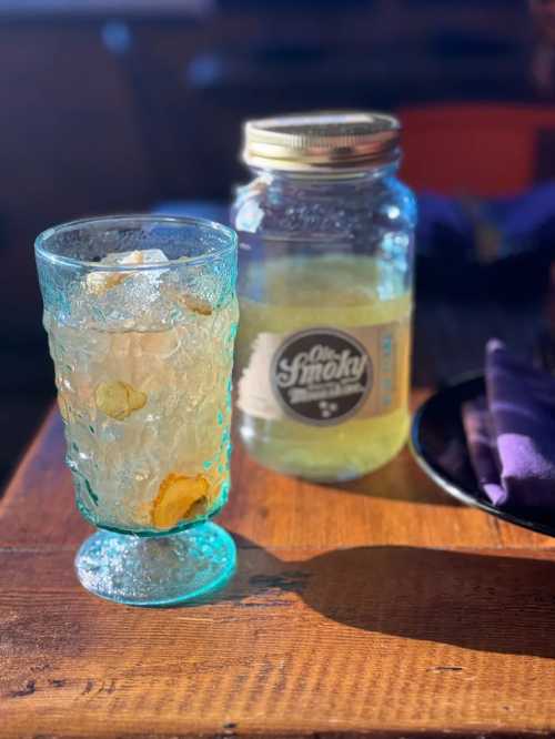 A glass of sparkling drink with ice sits next to a jar of Old Smoky on a wooden table.