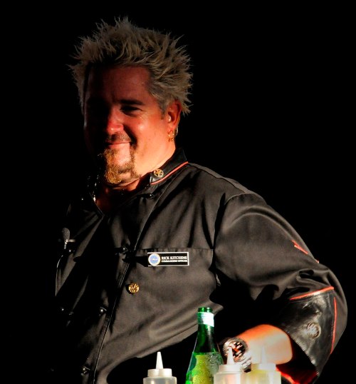 A chef with spiky blonde hair smiles while standing behind a counter with condiments and drinks.