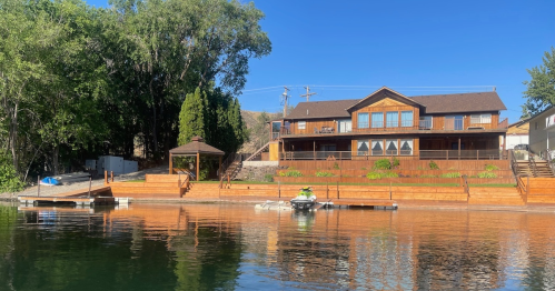 A lakeside house with a wooden deck, surrounded by trees, and a person on a jet ski near the dock. Clear blue sky.