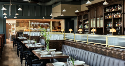 Elegant restaurant interior with wooden tables, leather seating, and a well-stocked bar in the background.