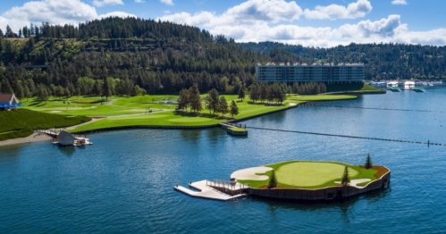 A scenic view of a golf course by a lake, surrounded by trees and a hotel in the background under a blue sky.